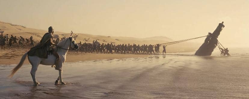 Seorang prajurit menunggang kuda di tepi pantai menyaksikan sekelompok tentara menarik kapal besar yang miring di pasir.