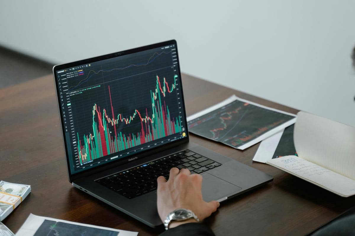 Seseorang menganalisis grafik pergerakan saham di layar laptop dengan data keuangan dan catatan di meja kerja kayu.