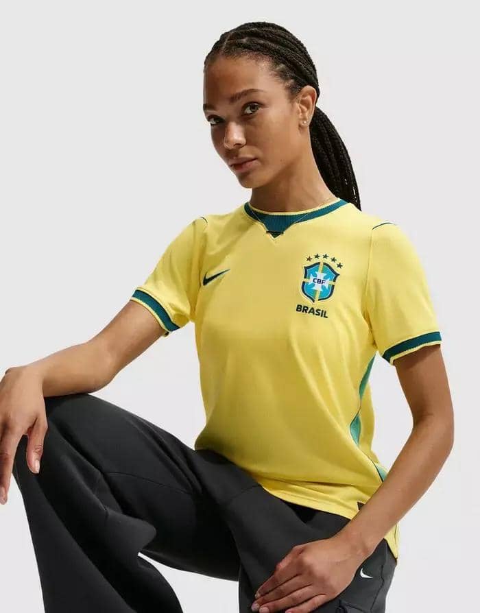 Seorang wanita mengenakan jersey tim nasional Brasil berwarna kuning dengan detail hijau, dipadukan dengan celana panjang hitam.
