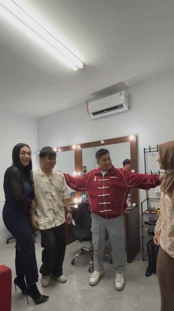 Empat orang berdiri di ruang rias, dua di antaranya tersenyum sambil berinteraksi akrab, dengan pencahayaan dari lampu di cermin.