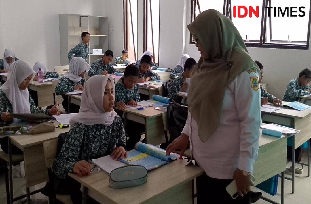 Guru perempuan berbicara dengan siswi di ruang kelas sekolah rakyat Banyumas, sementara siswa lain belajar di meja masing-masing.