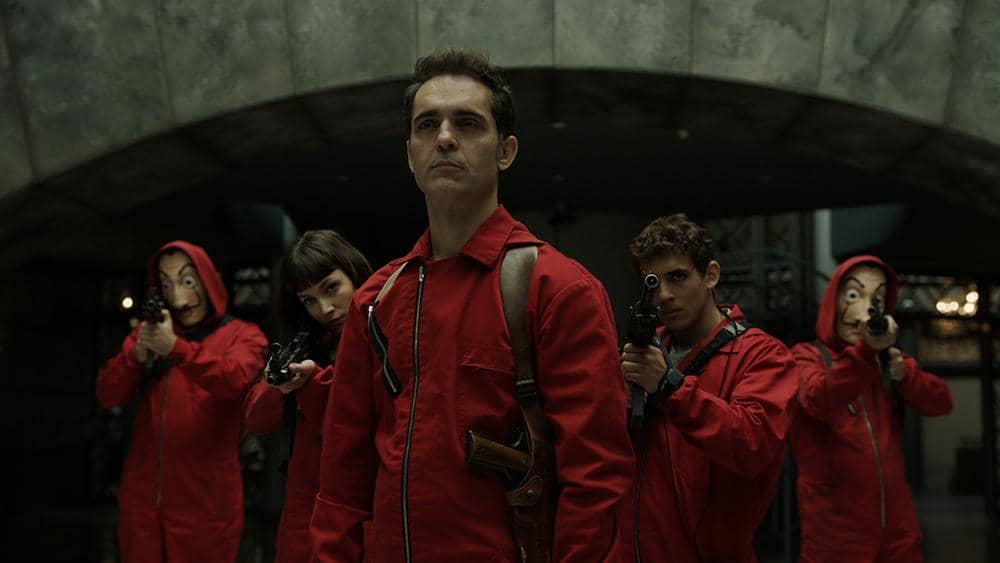 Lima perampok mengenakan jumpsuit merah dan membawa senjata api berdiri di bawah lengkungan bangunan dalam adegan serial Money Heist.