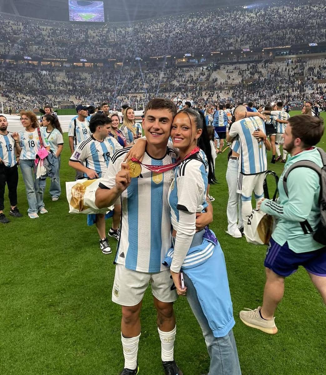 Seorang pemain sepak bola mengenakan jersey Argentina memegang medali emas sambil berpose dengan seorang wanita di stadion penuh penonton.