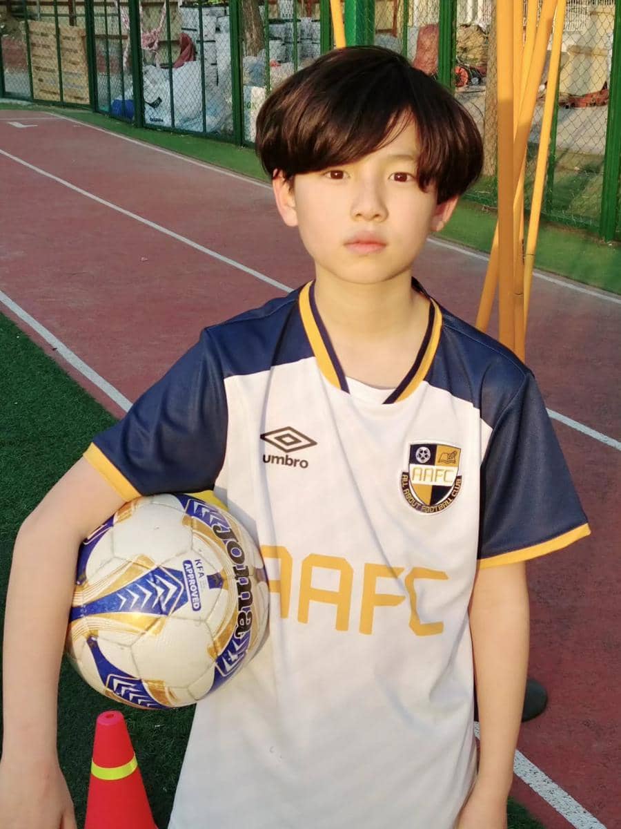 Seorang anak mengenakan seragam sepak bola AAFC berwarna putih dan biru sambil memegang bola di lapangan olahraga.