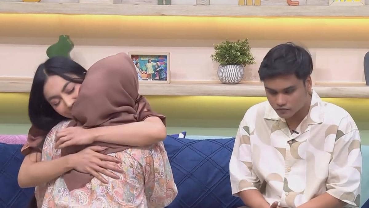 Seorang wanita berhijab memeluk wanita berambut hitam di ruang tamu, sementara seorang pria duduk di samping mereka dengan ekspresi serius.