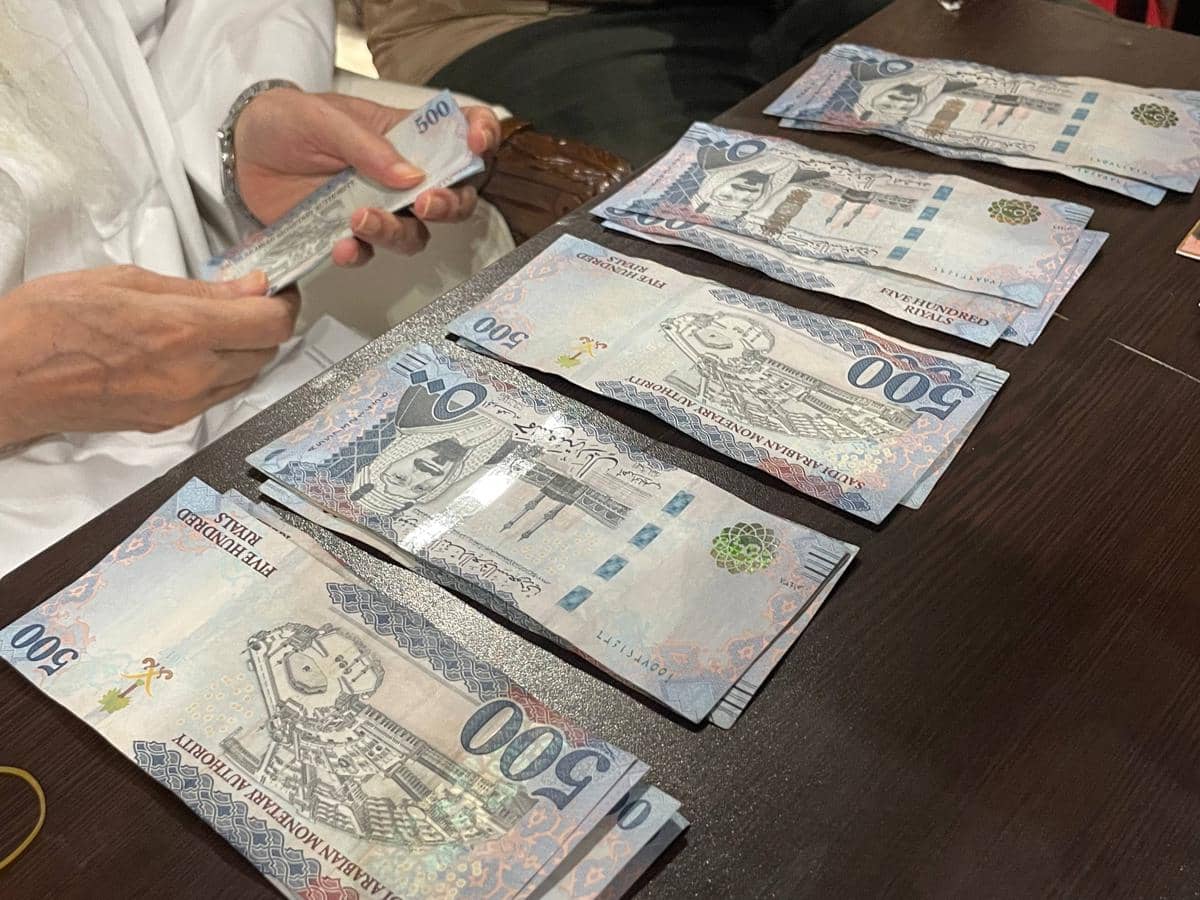 Seseorang menghitung lembaran uang kertas 500 riyal Arab Saudi yang tersusun rapi di atas meja kayu gelap.