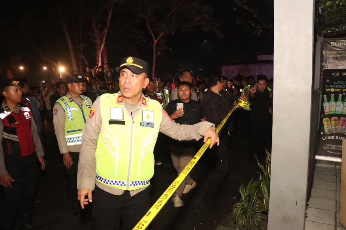 Petugas polisi memasang garis polisi di depan kafe pada malam hari dengan kerumunan warga yang menyaksikan di belakangnya.