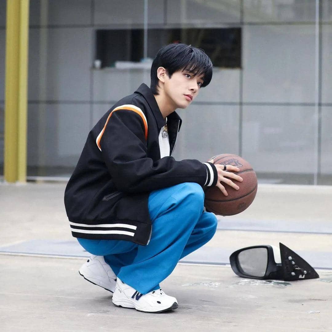 Seorang pria berambut hitam mengenakan jaket hitam dan celana biru jongkok sambil memegang bola basket di area luar ruangan.