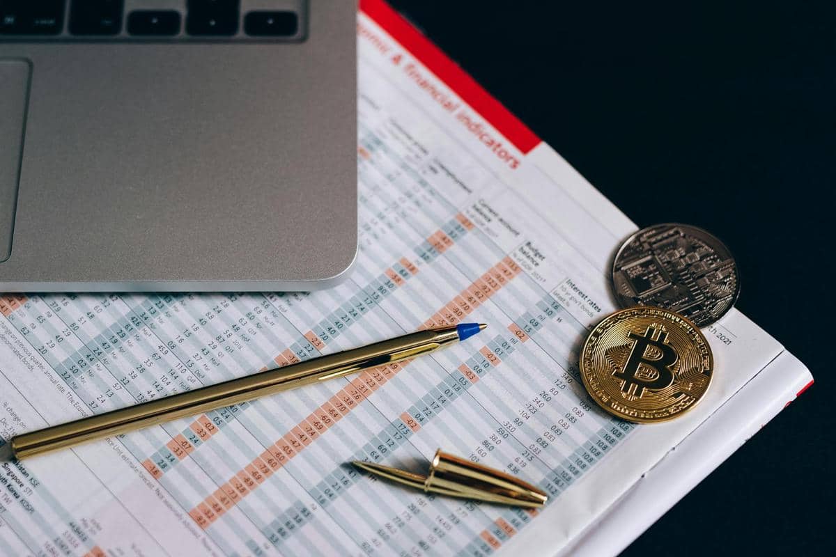 Koin kripto, pena emas, dan laptop di atas lembar data keuangan yang mencerminkan konsep ekonomi digital modern.