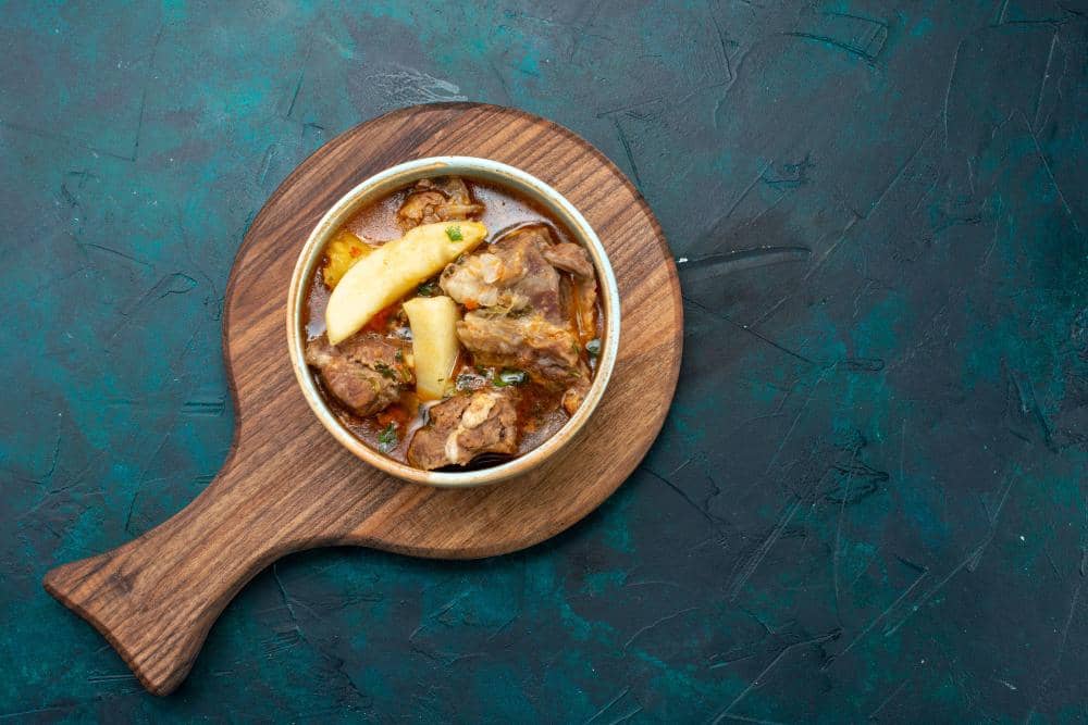 Semangkuk sop iga sapi dengan potongan kentang disajikan di atas talenan kayu di atas meja berwarna gelap.