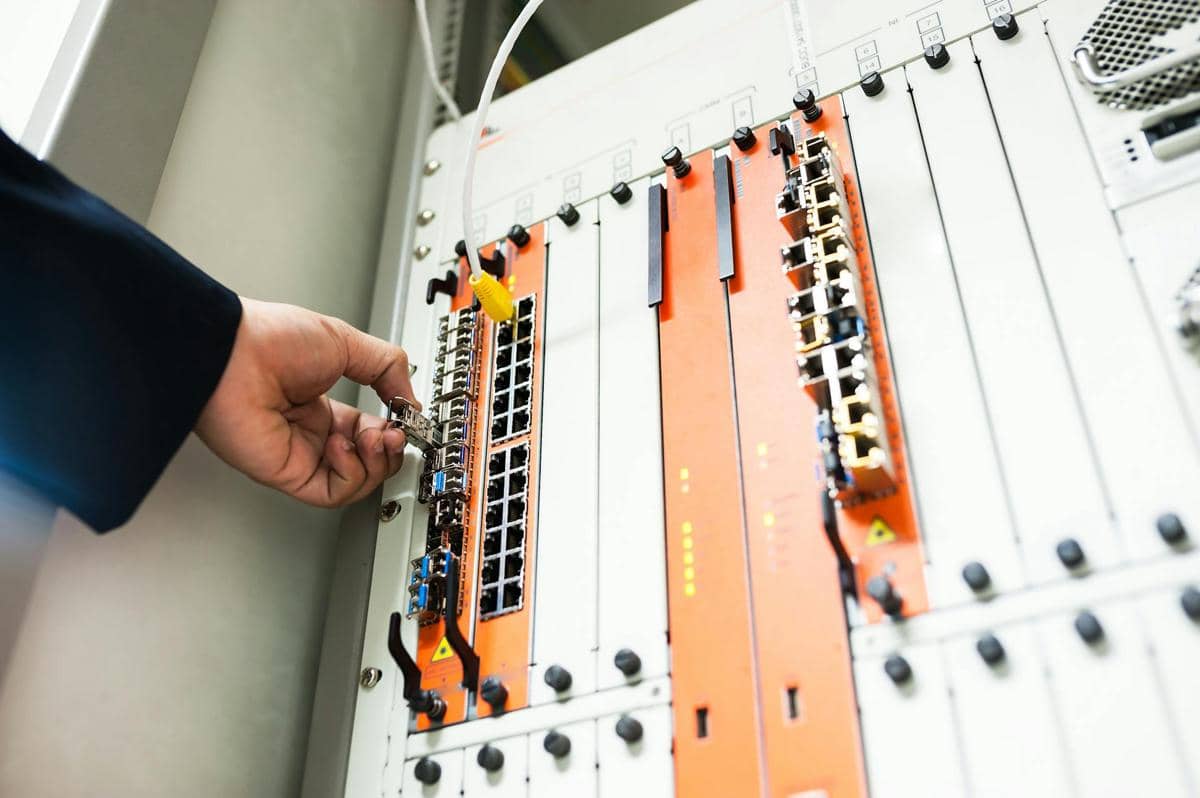 Tangan teknisi menghubungkan kabel optik ke panel jaringan dengan port dan indikator lampu di ruang server.