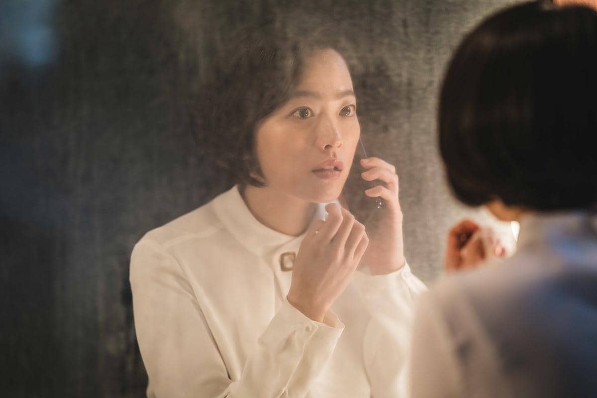 Seorang wanita berambut pendek mengenakan kemeja putih menatap cermin dengan ekspresi tegang sambil memegang ponsel di tangannya.