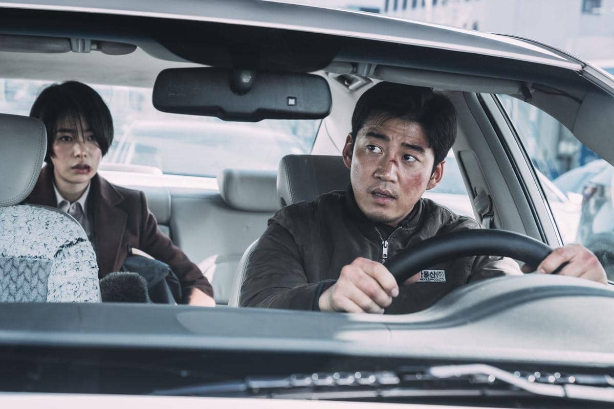 Seorang pria dengan wajah terluka duduk di kursi pengemudi mobil sementara seorang wanita duduk di kursi penumpang belakang dengan ekspresi tegang.