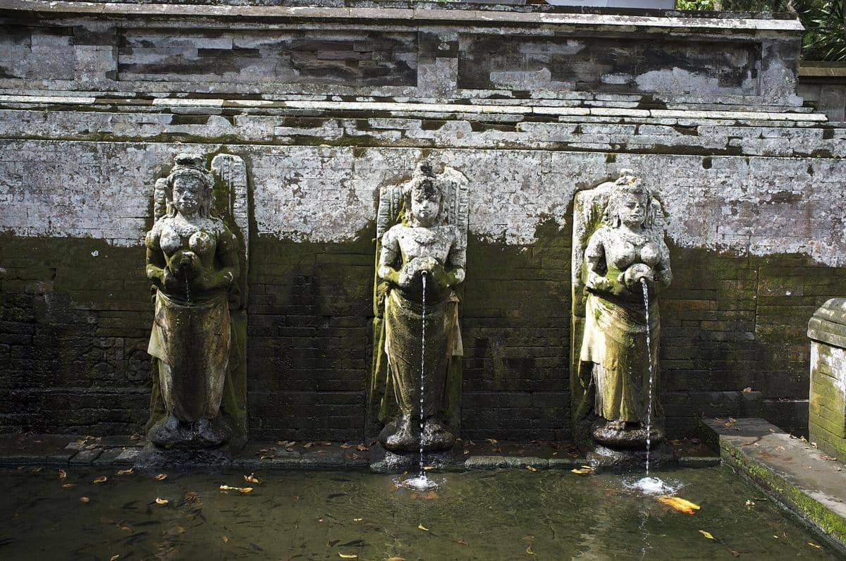 Tiga patung batu berlumut memancurkan air ke kolam di dalam kompleks Goa Gajah, dikelilingi dinding batu tua berlumut.