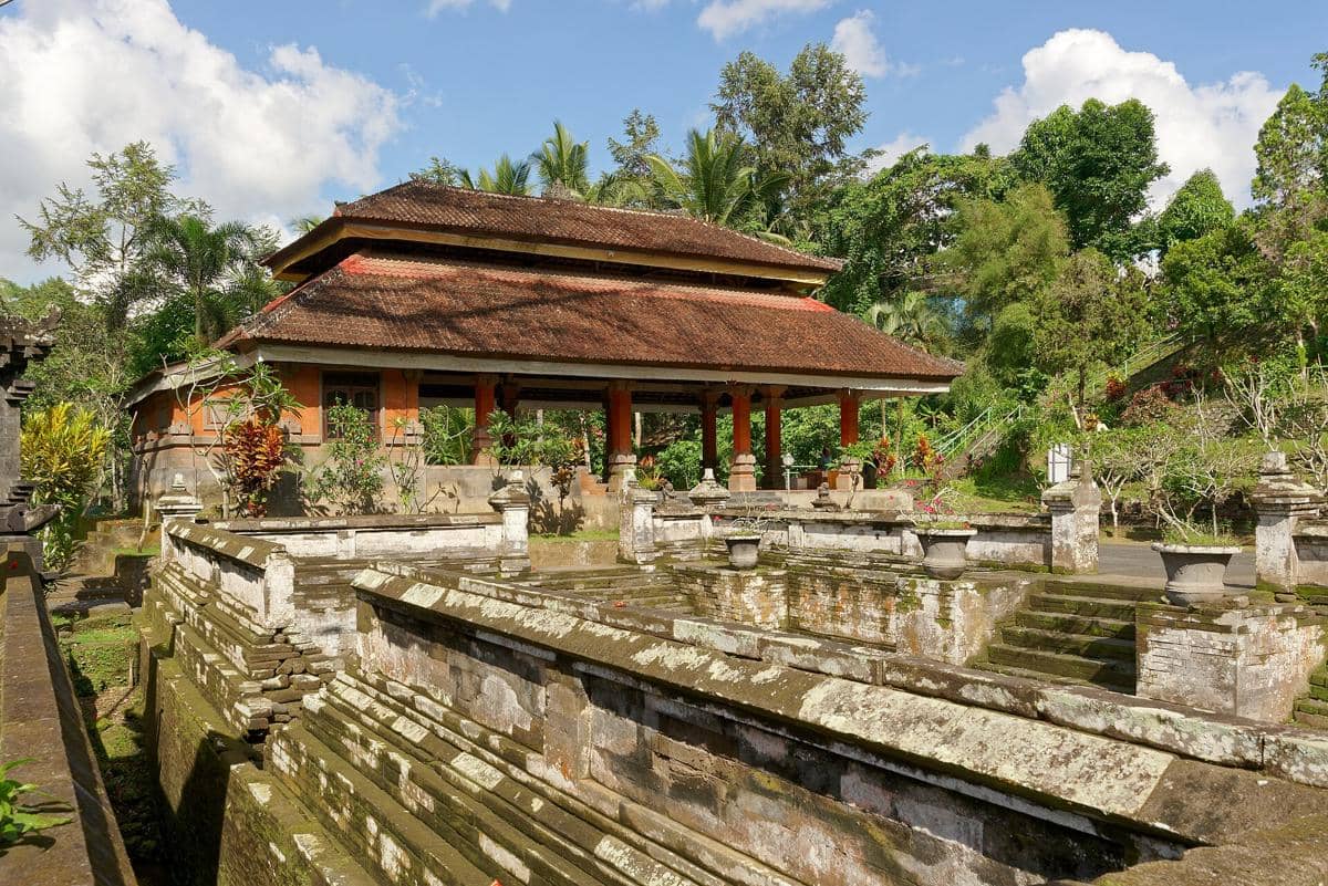 Bangunan beratap tradisional dengan dinding bata merah dan kolam batu berlumut di kompleks Goa Gajah, dikelilingi pepohonan hijau.
