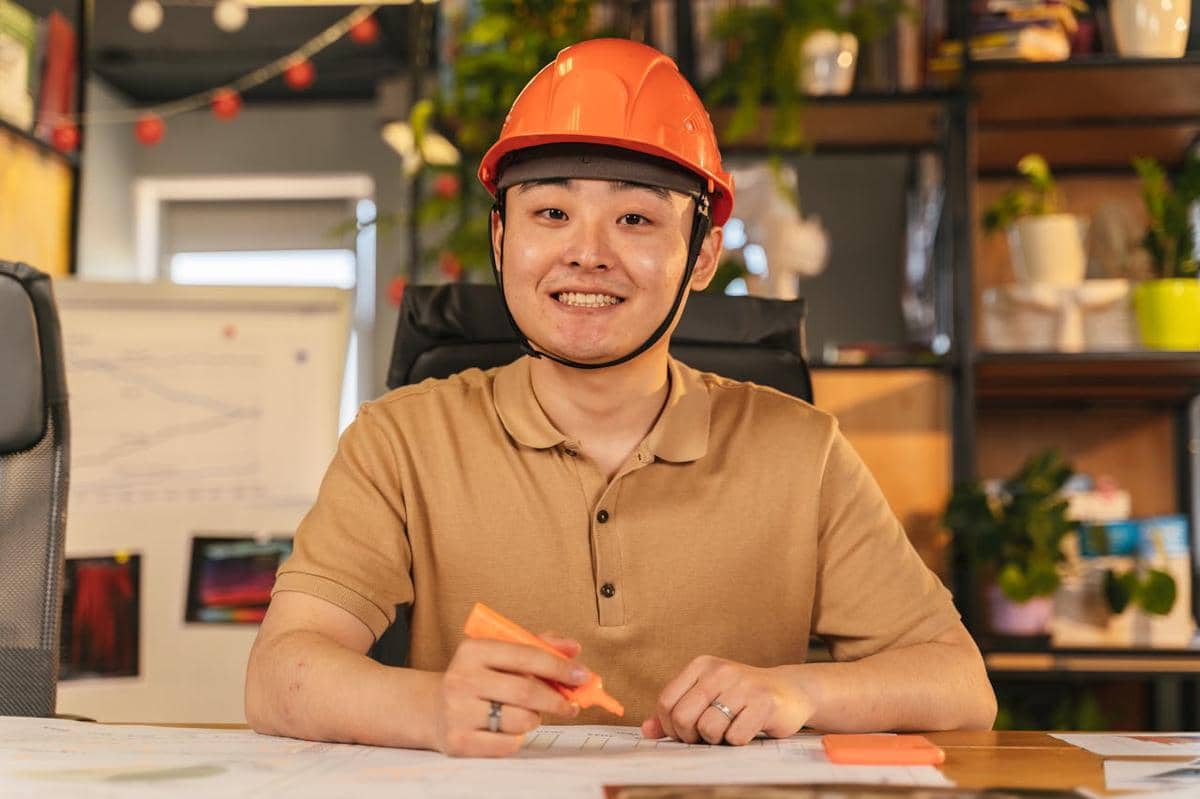 Seorang pekerja mengenakan helm oranye tersenyum sambil memegang stabilo di meja kerja dengan latar belakang kantor yang hangat.