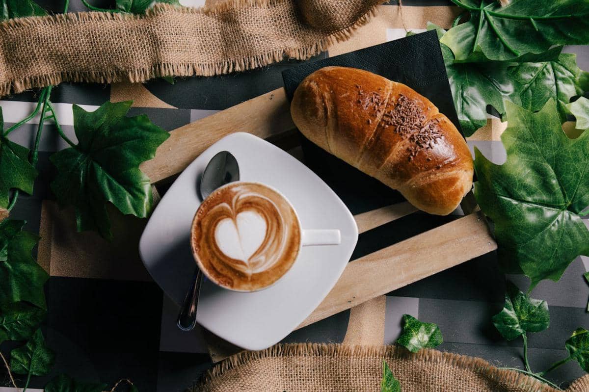 Secangkir kopi dengan latte art berbentuk hati disajikan bersama roti salt bread di atas meja dengan hiasan daun hijau.