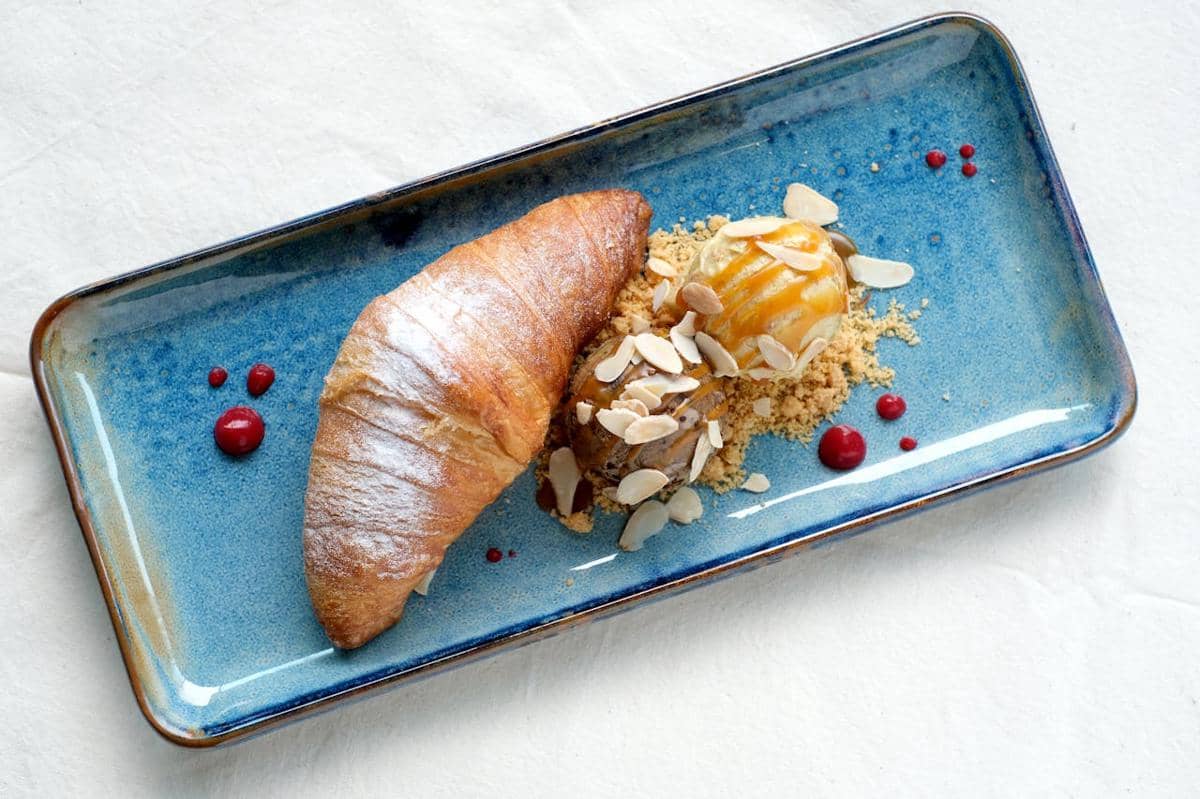 Roti salt bread disajikan di piring biru bersama es krim dengan saus karamel, taburan kacang almond, dan remah biskuit.