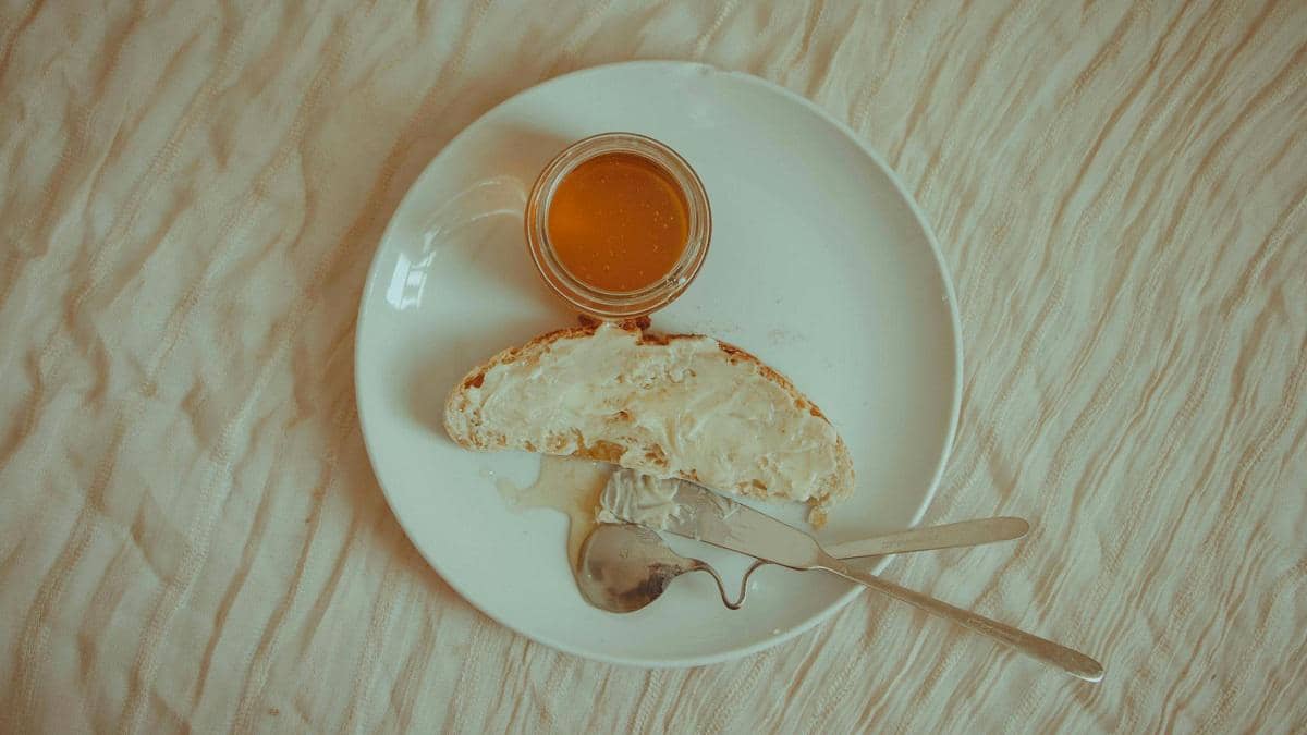 Sepotong roti asin dengan olesan mentega di atas piring putih, disajikan bersama madu dalam toples kecil dan sendok di sampingnya.
