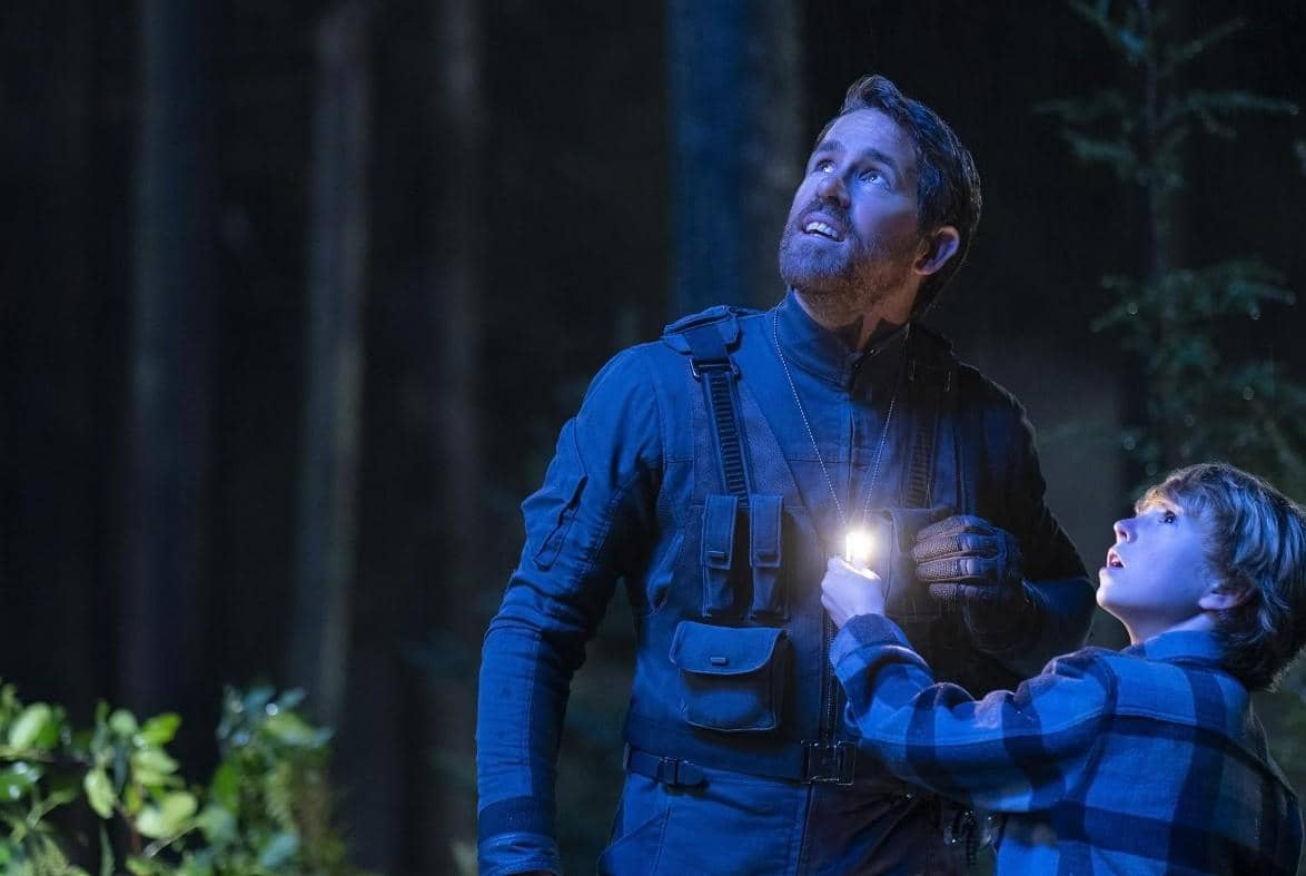 Dua karakter dalam film The Adam Project berdiri di hutan malam hari, melihat ke atas dengan cahaya kecil di antara mereka.