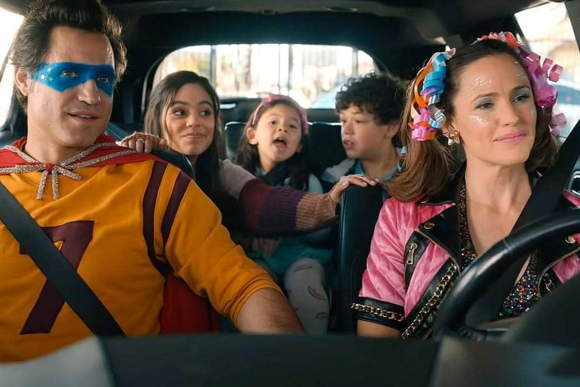 Adegan keluarga di dalam mobil dari film Yes Day, menampilkan orang tua dan tiga anak yang tampak ceria dengan kostum berwarna-warni.