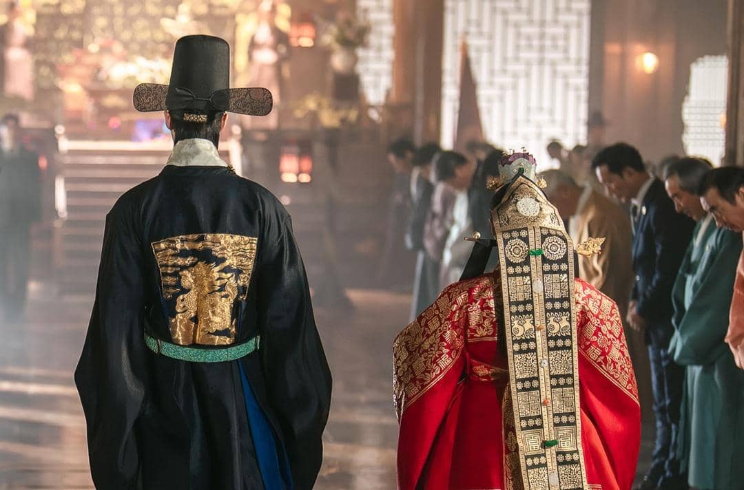 Dua tokoh mengenakan hanbok kerajaan tradisional Korea berdiri di aula istana dengan para pejabat membungkuk di sekeliling mereka.