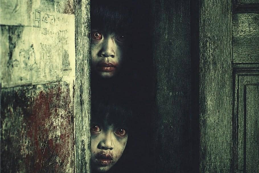 Dua anak berwajah pucat dengan mata merah menatap dari balik pintu kayu tua dalam suasana gelap dan menyeramkan.