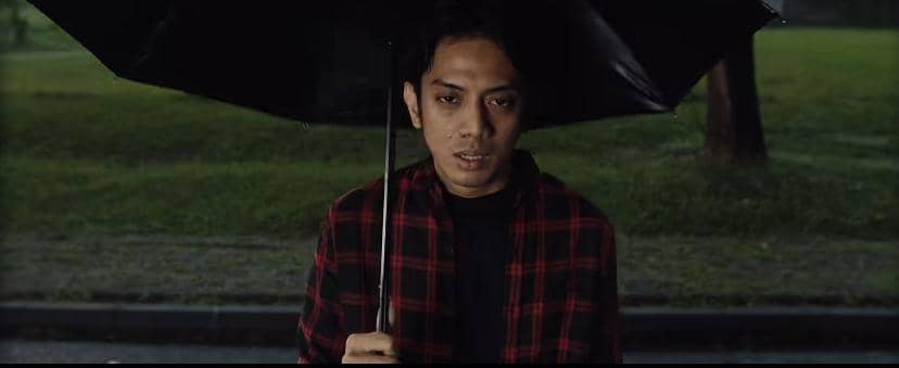 Seorang pria mengenakan kemeja kotak-kotak merah hitam memegang payung hitam di luar ruangan dengan latar taman hijau saat suasana mendung.