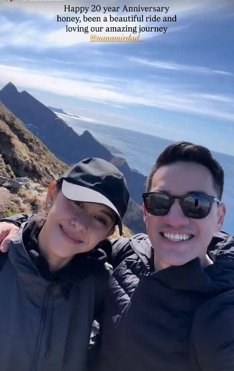 Sepasang suami istri tersenyum sambil berpose di pegunungan Norwegia dengan latar laut dan langit cerah di belakang mereka.