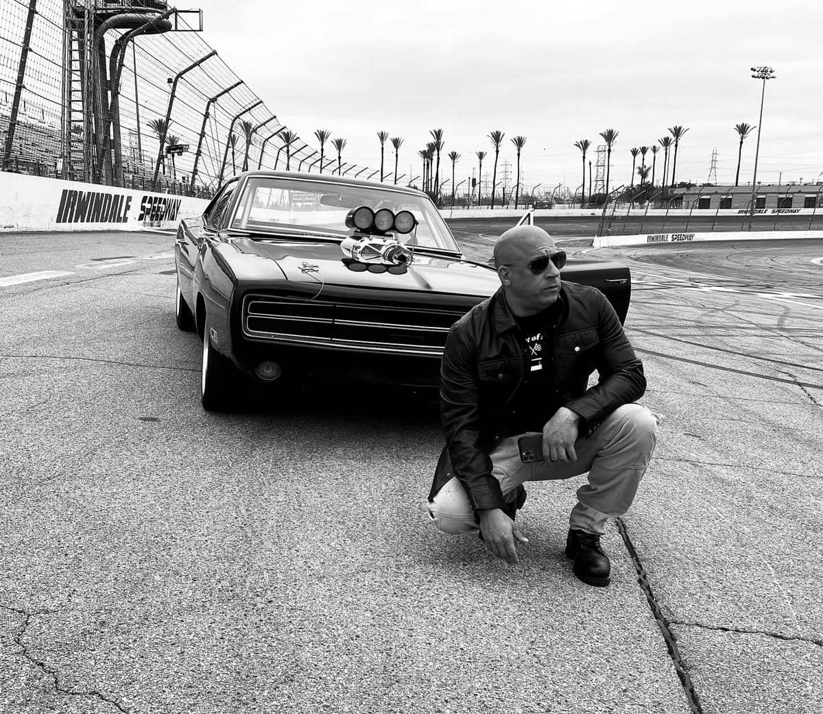 Seorang pria berkacamata hitam berpose jongkok di depan mobil klasik berwarna hitam di lintasan balap Irwindale Speedway.