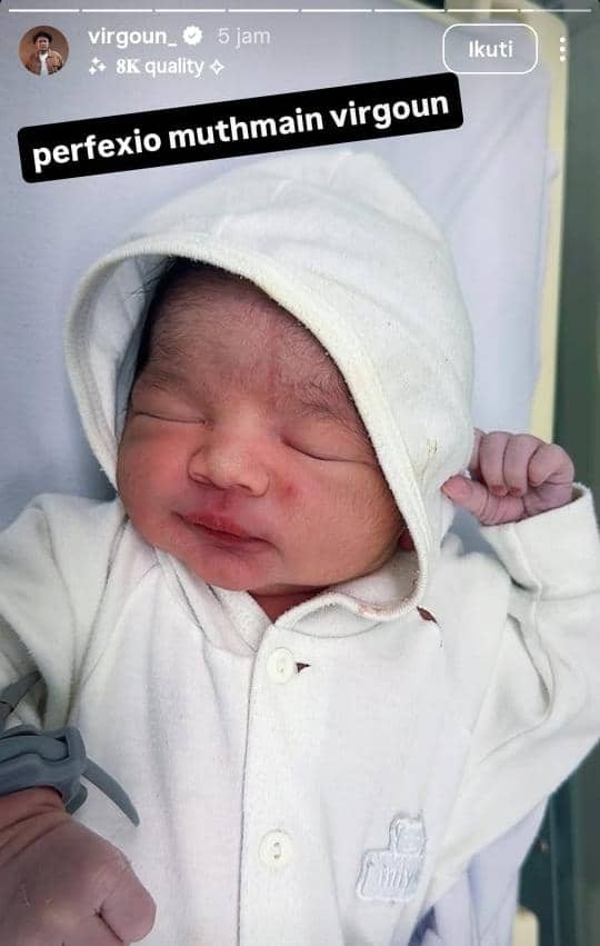 Bayi baru lahir mengenakan pakaian dan hoodie putih sedang berbaring di tempat tidur rumah sakit dengan tangan menggenggam lembut.