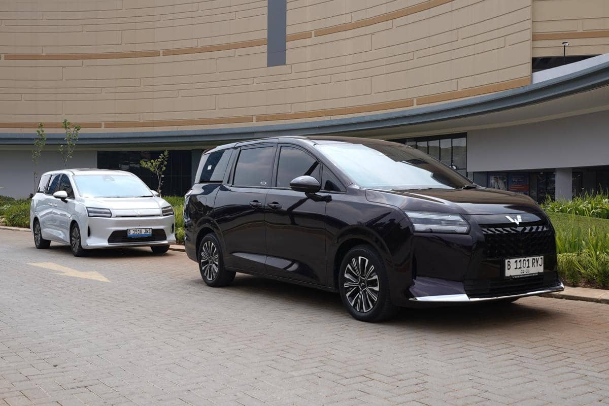 Dua mobil Wuling berwarna hitam dan putih terparkir di area luar gedung dengan latar dinding melengkung berwarna krem.
