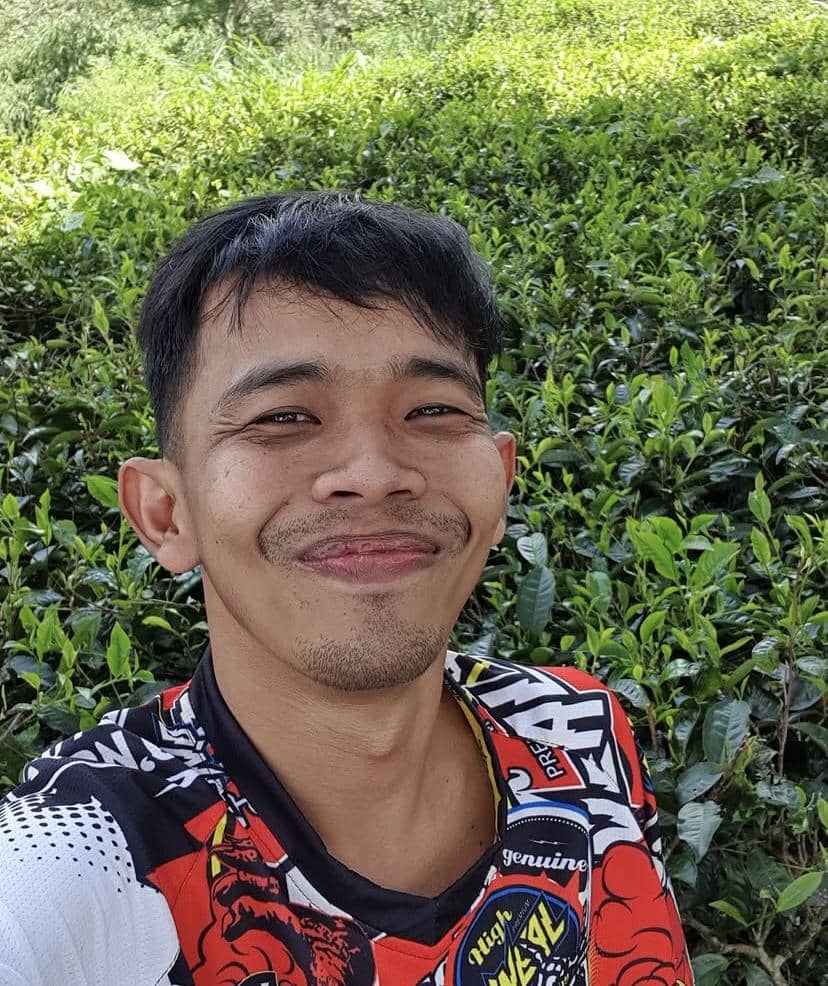 Seorang pria tersenyum mengenakan kaus berwarna merah, hitam, dan putih dengan latar belakang tanaman hijau yang rimbun.