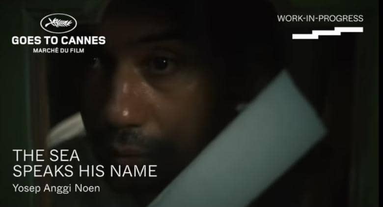 Seorang pria terlihat dari dekat dalam pencahayaan redup dengan teks promosi film 'The Sea Speaks His Name' karya Yosep Anggi Noen di Marche du Film Cannes.