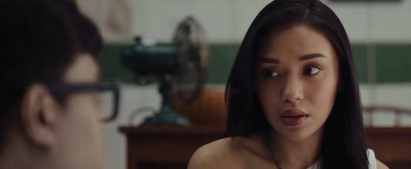 Seorang perempuan berambut panjang menatap seseorang di depannya di sebuah ruangan dengan kipas angin dan meja kayu di latar belakang.