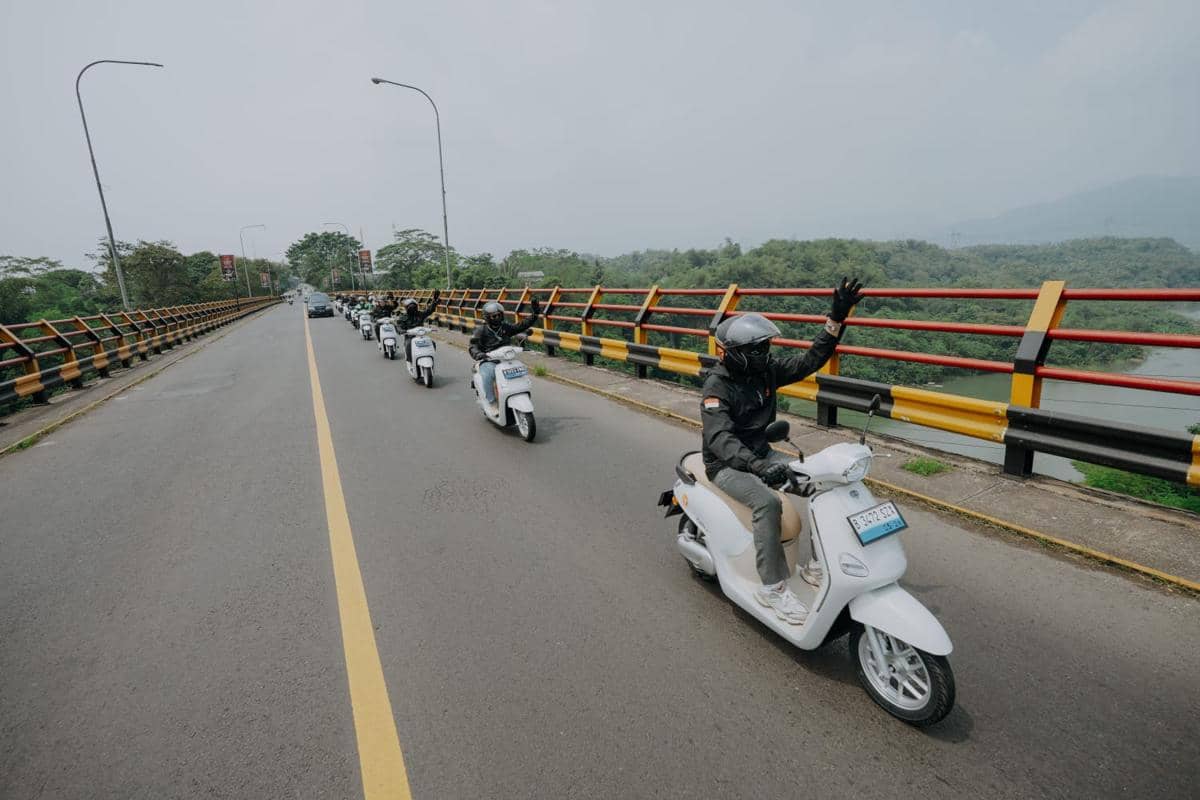 Rombongan pengendara skuter putih melintasi jembatan dengan pemandangan hijau di sekitarnya, salah satu pengendara melambaikan tangan.