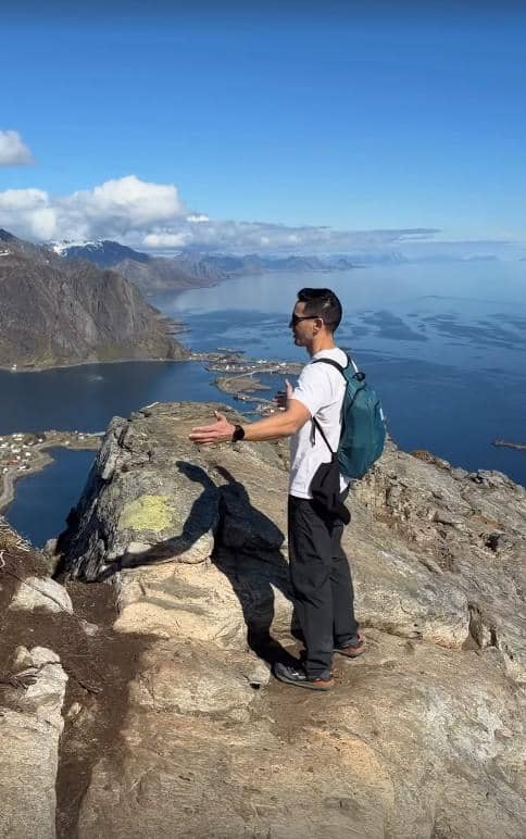 Seorang pria berdiri di tepi tebing berbatu dengan pemandangan laut dan pegunungan di Norwegia di bawah langit cerah.