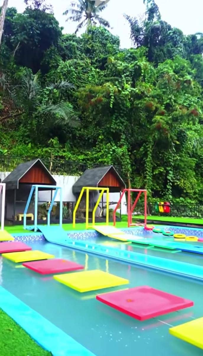 Area permainan air berwarna-warni di The Nice Park Lampung dengan latar pepohonan hijau dan rumah kecil beratap hitam.
