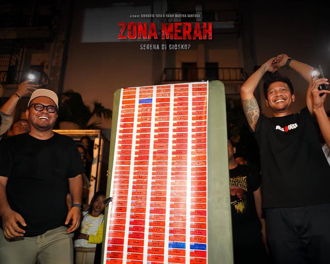 Dua pria tersenyum di depan papan berisi deretan kartu merah dengan tulisan Zona Merah di atasnya, diambil pada malam hari.
