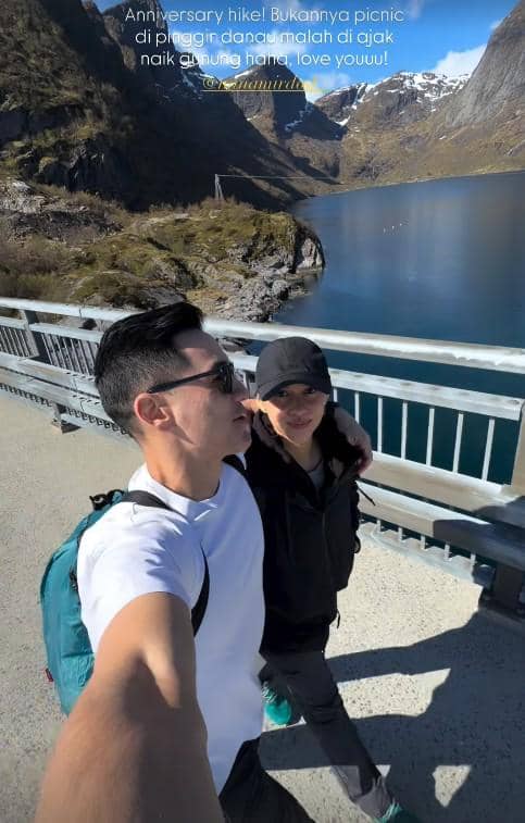 Sepasang wisatawan berjalan di jembatan dengan latar pegunungan bersalju dan danau jernih di Norwegia saat cuaca cerah.