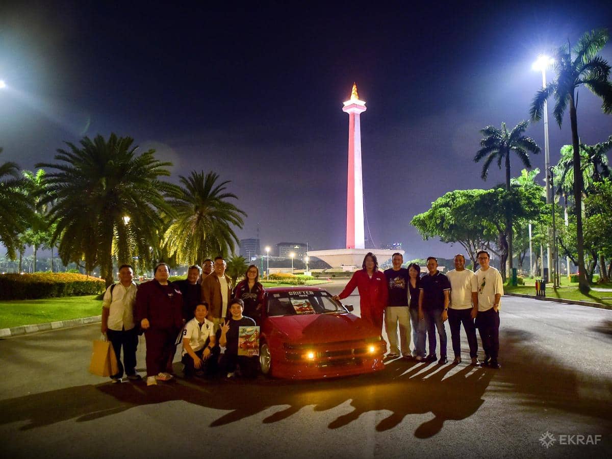Sekelompok orang berpose bersama mobil sport merah di depan Monas yang bercahaya pada malam hari di Jakarta.