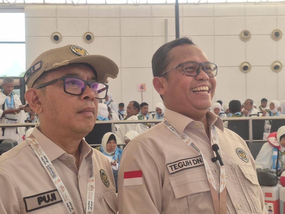 Dua pejabat Kementerian Agama mengenakan seragam krem dengan tanda pengenal di Bandara King Abdul Aziz Jeddah, dikelilingi jamaah haji.
