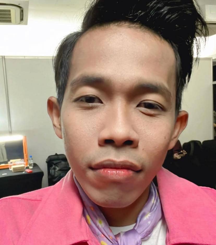Seorang pria mengenakan jaket merah muda dan syal ungu berpose di dalam ruangan dengan meja rias di latar belakang.