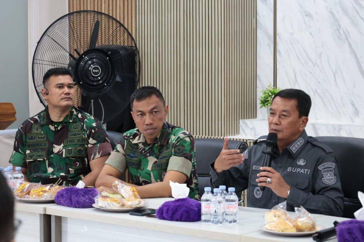 Bupati Tangerang berbicara memegang mikrofon didampingi dua anggota TNI dalam pertemuan di ruang rapat dengan hidangan di atas meja.