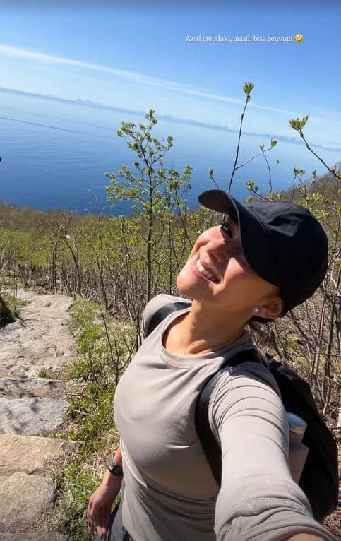 Seorang perempuan tersenyum sambil berfoto selfie di jalur pendakian dengan latar laut biru dan pepohonan hijau di Norwegia.