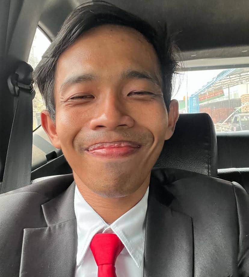 Seorang pria mengenakan jas hitam, kemeja putih, dan dasi merah tersenyum saat duduk di dalam mobil dengan sabuk pengaman terpasang.