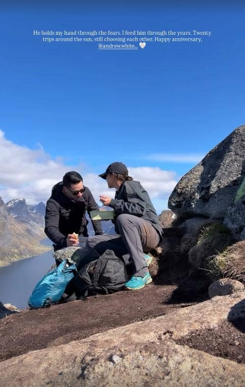 Sepasang wisatawan duduk di tepi tebing berbatu di Norwegia, menikmati pemandangan pegunungan dan danau di bawah langit cerah.