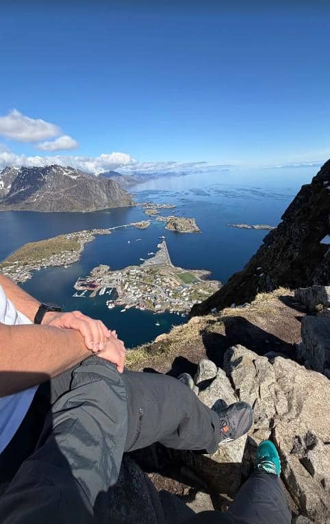 Dua orang duduk di tepi tebing dengan pemandangan laut, pulau kecil, dan pegunungan bersalju di Norwegia di bawah langit cerah.