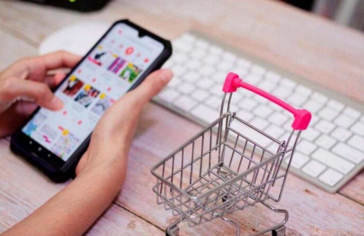 “Seseorang menggunakan smartphone untuk berbelanja online dengan miniatur keranjang belanja di atas meja kerja.”
