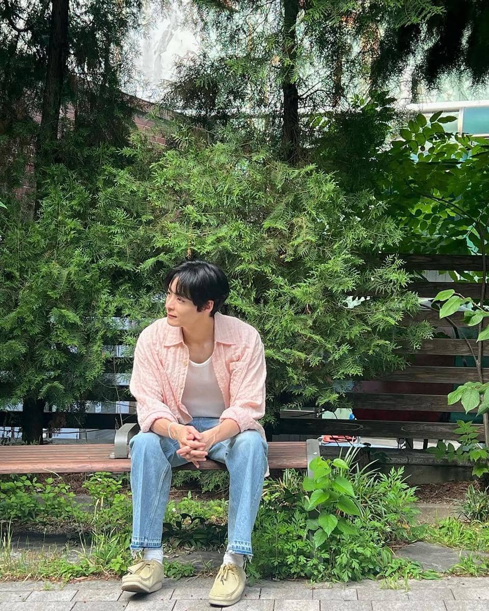 Pria duduk di bangku taman mengenakan kemeja merah muda, kaus putih, celana jeans biru, dan sepatu krem dengan latar pepohonan hijau.